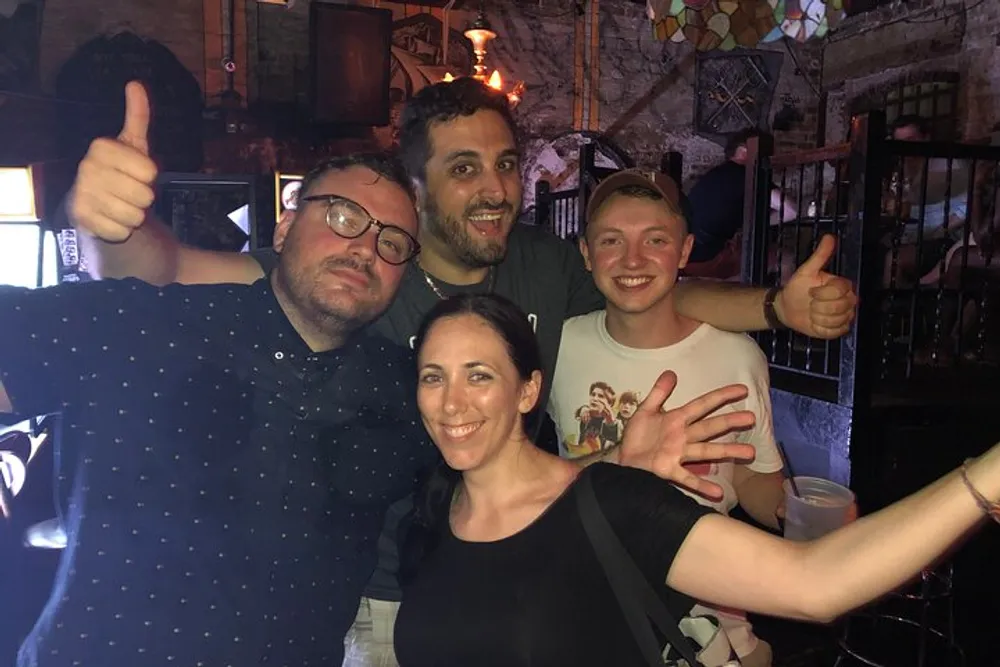 Four friends are posing for a group photo with smiles and thumbs-up gestures seemingly enjoying a good time together in a bar-like setting