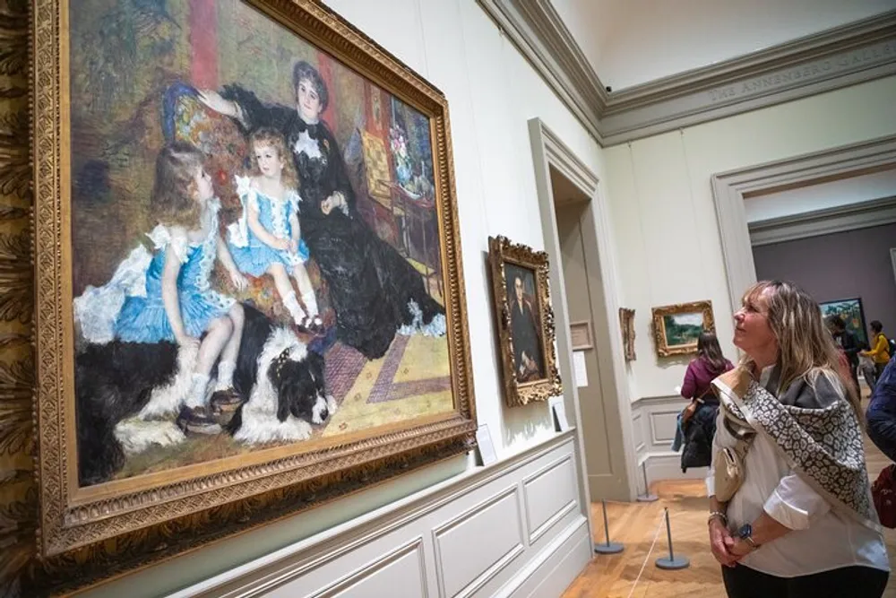 A person is admiring a large Impressionist painting featuring two children and a dog displayed in an ornate gold frame in a gallery with other artworks