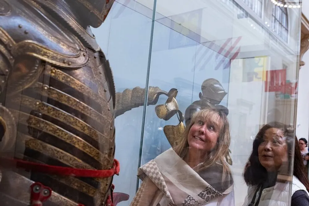 Two women are looking at a suit of armor on display through a glass case with one womans reflection aligning with the helmet making it look like she is wearing it