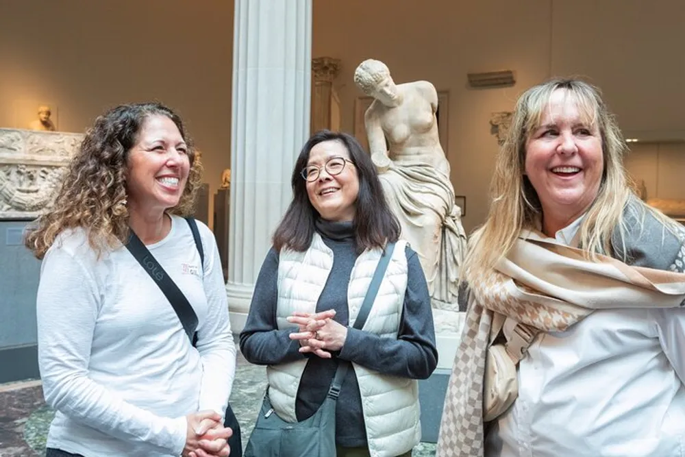 Three smiling women stand in front of a classical statue in what appears to be a museum setting