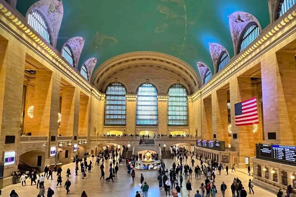 The photo shows the bustling interior of Grand Central Terminal with people milling about under the expansive arched windows and a large American flag hanging prominently