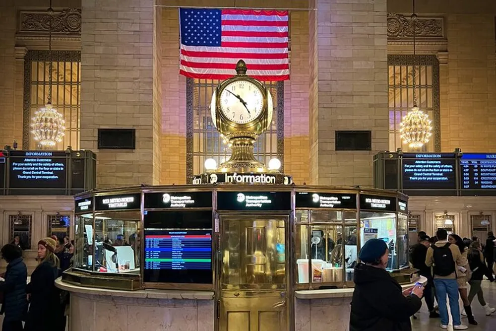 The image shows the iconic information booth with its opal-faced clock in Grand Central Terminal New York below a large American flag with people milling around in the busy transportation hub