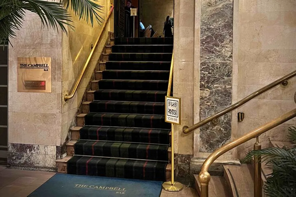 This image shows a carpeted staircase leading to The Campbell Bar with brass railings a SCAN ME QR code sign and a person partially visible at the top of the stairs