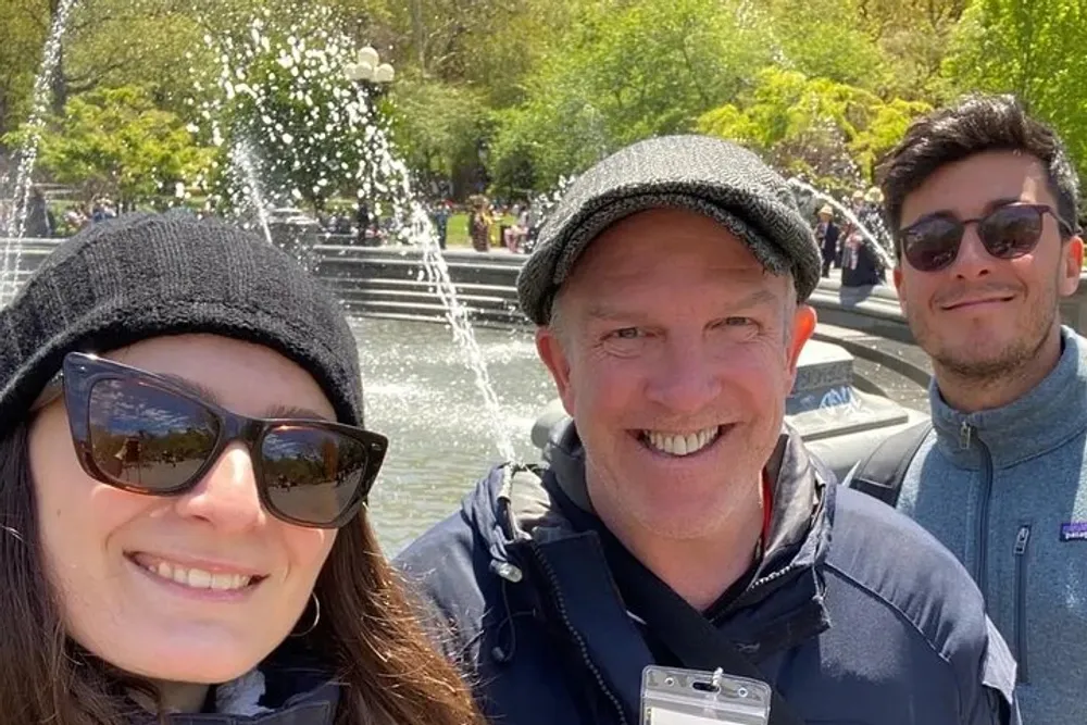 Three smiling people are taking a group selfie with a fountain in the background on a sunny day