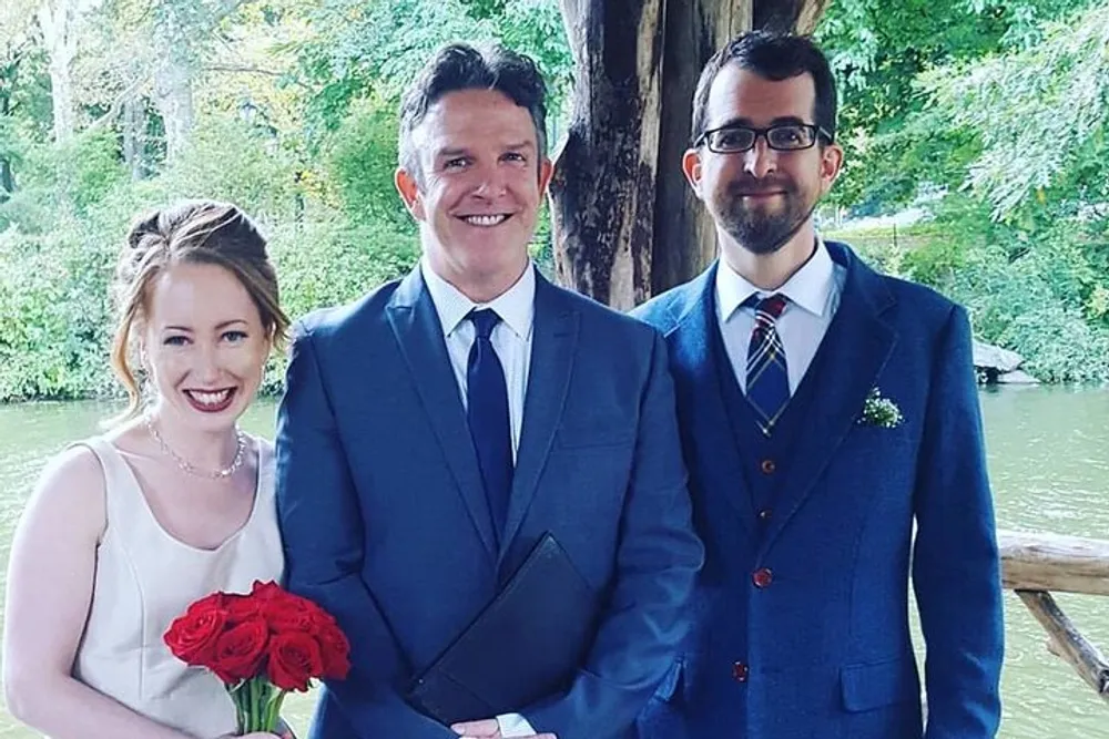 Three people are dressed up for a formal occasion standing outdoors with a background of greenery and a body of water with the woman holding a bouquet of red roses