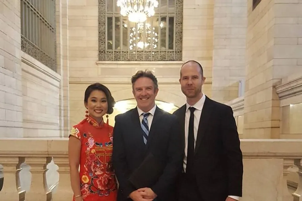 Three people are smiling for the camera in an elegant hallway with a chandelier in the background the person on the left is wearing a red traditional dress while the two men are in suits