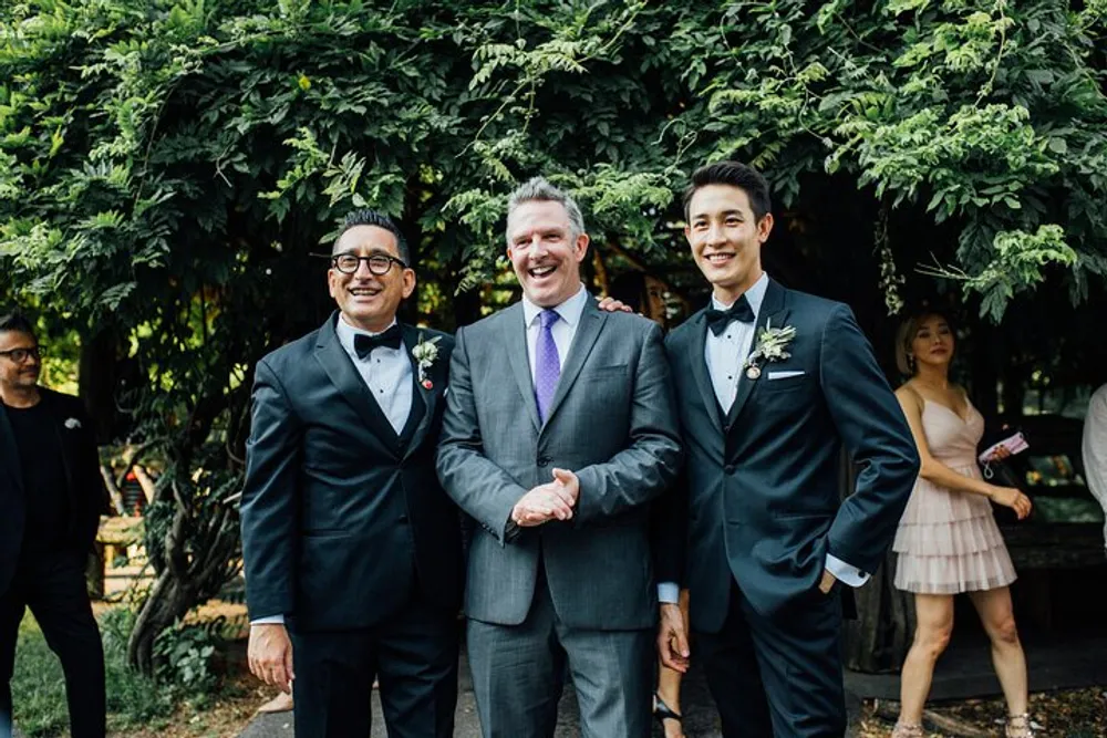 Three smiling men in formal suits are posing for a photo likely at a wedding given the boutonnieres and festive attire