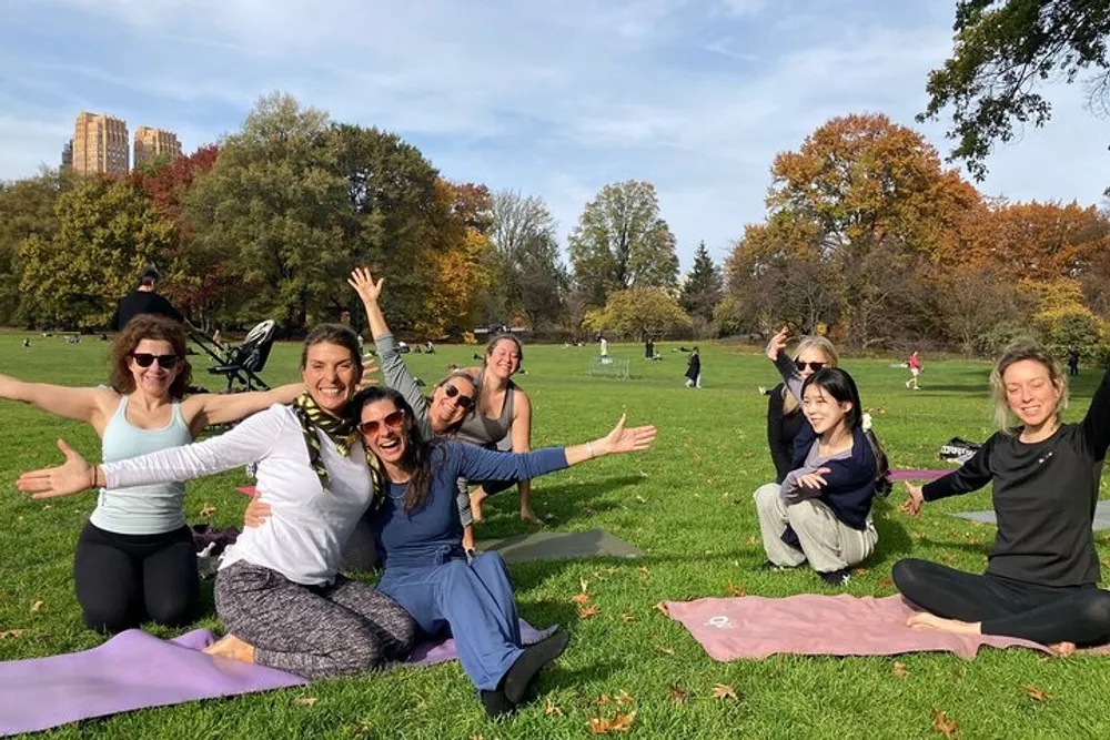 A group of people is smiling and posing playfully on yoga mats in a sunny park with autumn foliage in the background