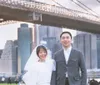 A happy couple in formal attire stands in front of the Brooklyn Bridge with the New York City skyline in the background