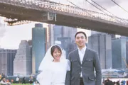 A happy couple in formal attire stands in front of the Brooklyn Bridge with the New York City skyline in the background.