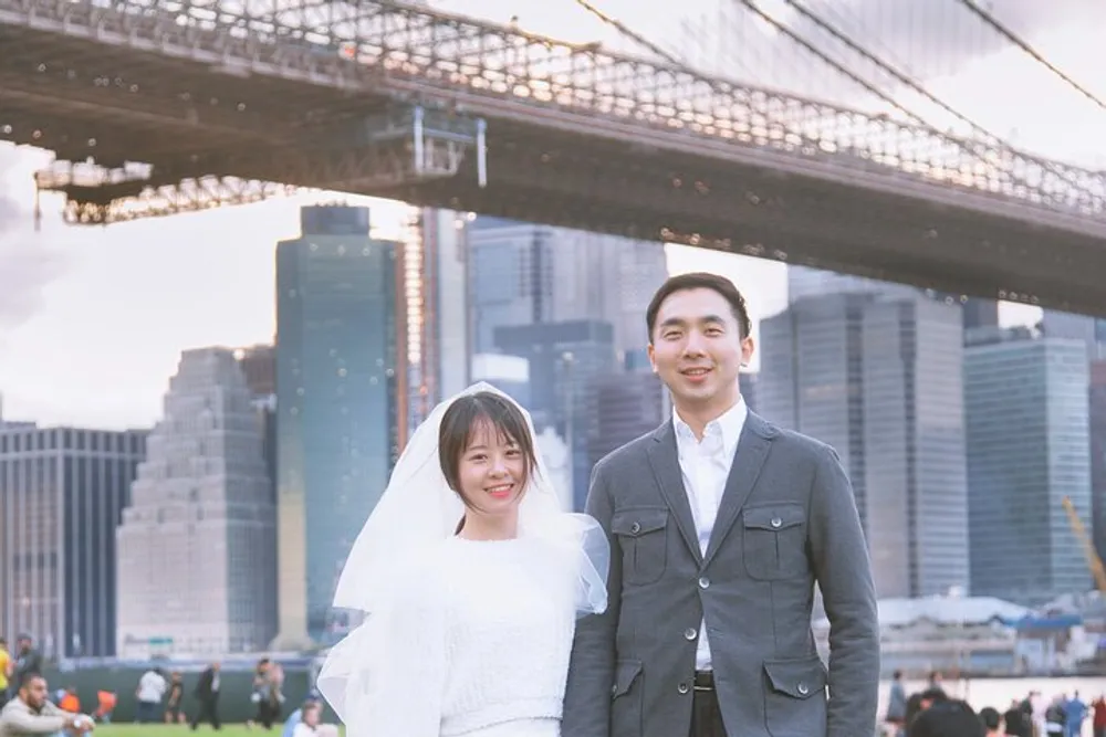 A happy couple in formal attire stands in front of the Brooklyn Bridge with the New York City skyline in the background