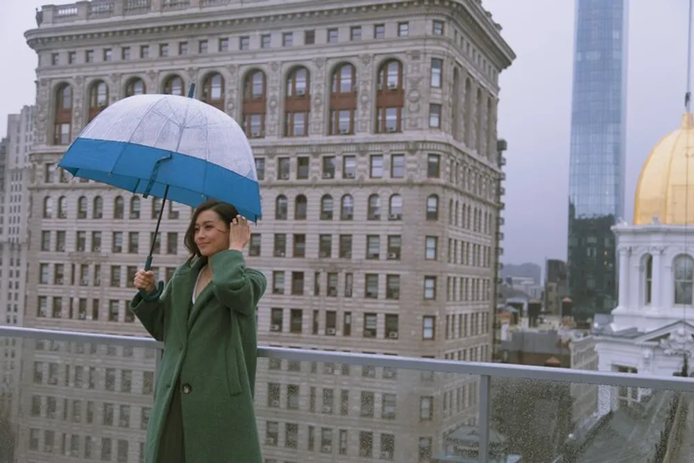 A person is standing on a balcony with a patterned umbrella in front of a cityscape that features a flatiron building on an overcast day