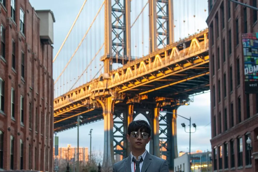 A person wearing sunglasses and a suit stands in the foreground with an illuminated suspension bridge and brick buildings in the background during what appears to be the golden hour