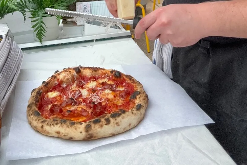 A person is grating cheese onto a freshly baked pizza with a golden-brown crust