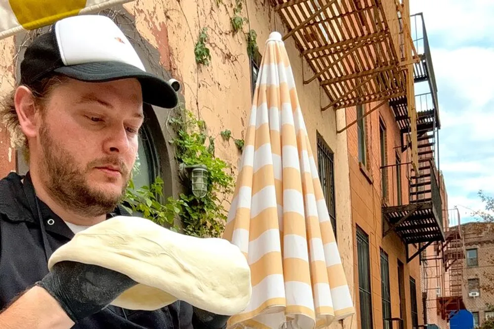 A person is focused on shaping a large piece of dough outdoors with a building and a striped umbrella in the background