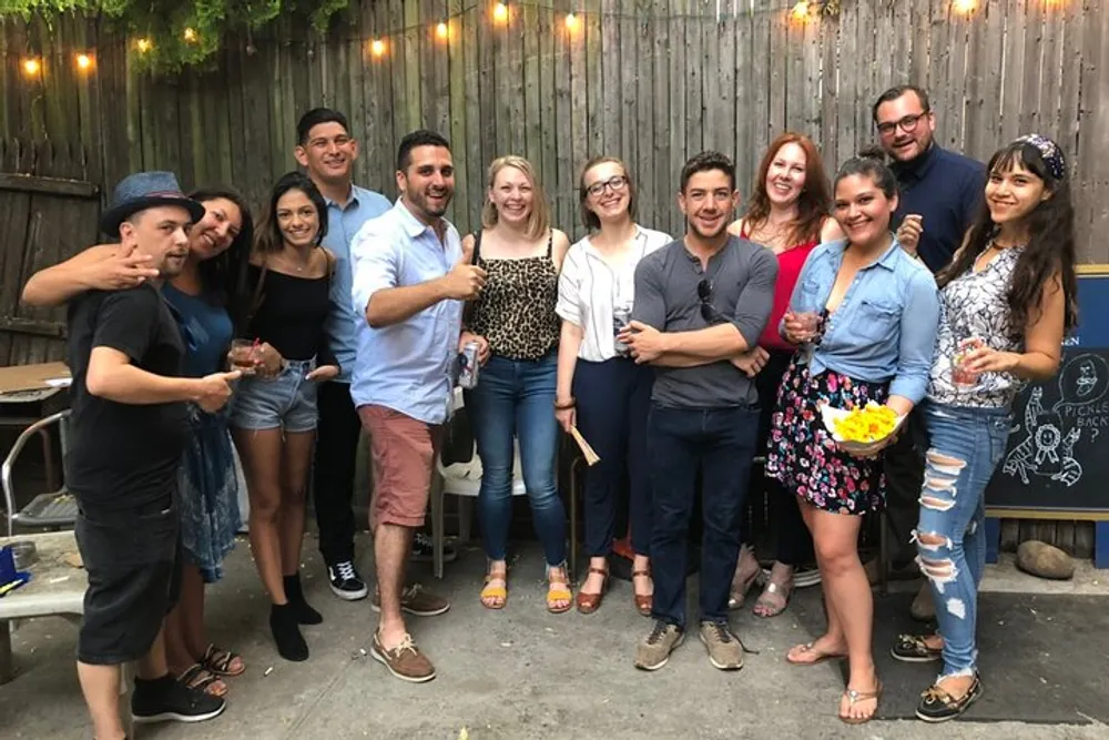 A group of smiling people are posing for a photo at an outdoor gathering some holding drinks and snacks suggesting a social event or celebration