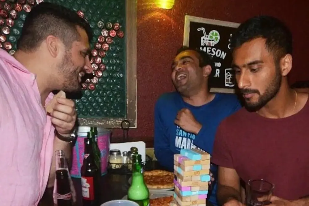 Three men are enjoying a fun moment with one laughing heartily and another concentrating on a game of Jenga at a table with pizza and drinks