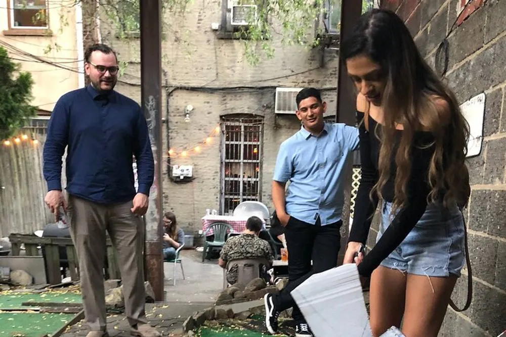 A group of people are socializing in an outdoor alleyway setting with a woman in the foreground seemingly adjusting her shoe or picking something up