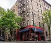 The image shows an urban street corner with a multi-story residential building a ground-floor restaurant with a red facade and tree-lined sidewalks evoking a typical New York City neighborhood vibe