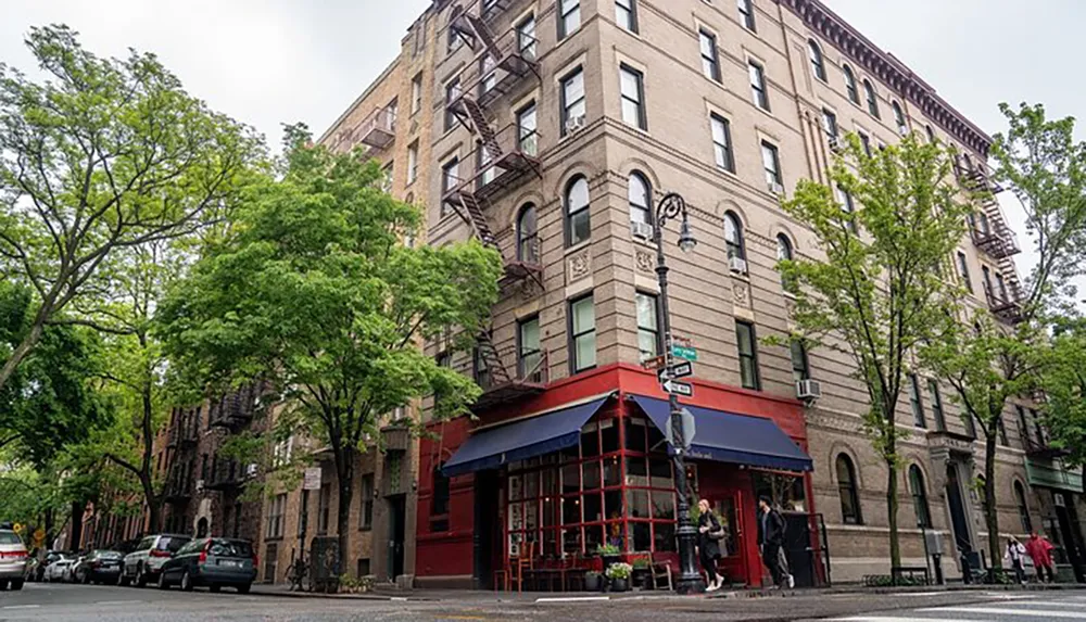 The image shows an urban street corner with a multi-story residential building a ground-floor restaurant with a red facade and tree-lined sidewalks evoking a typical New York City neighborhood vibe