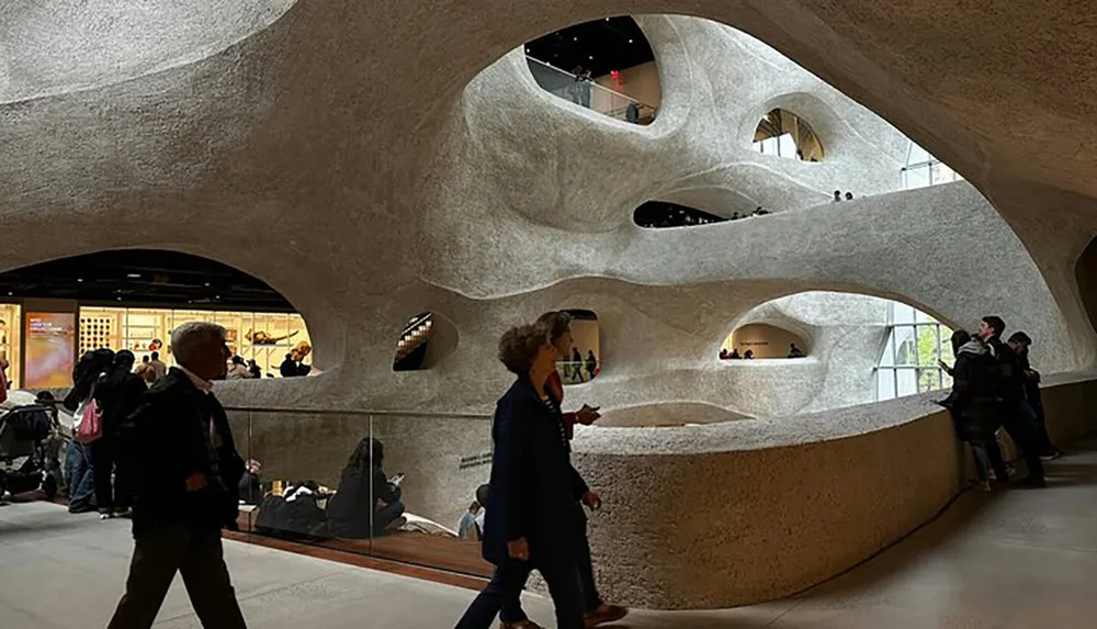 This image shows people walking through an organically designed space featuring curved walls with multiple rounded openings creating a sculptural cave-like atmosphere