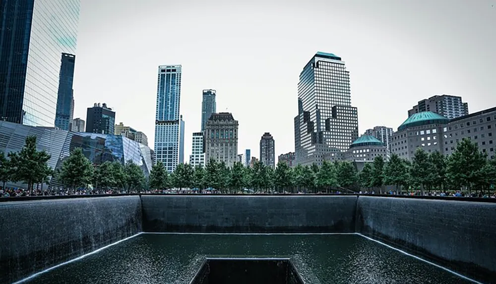 The image shows a serene reflection pool surrounded by trees and modern skyscrapers likely a memorial or urban park in a bustling city