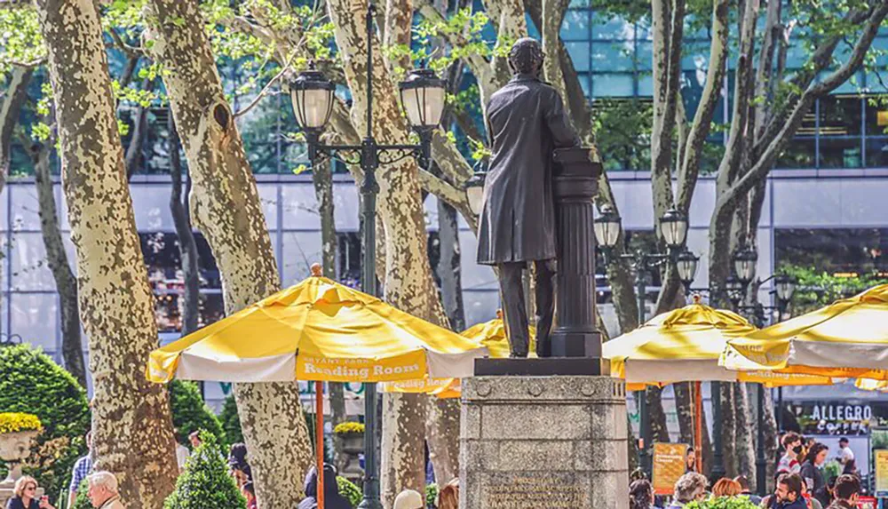 A statue stands overlooking a lively urban park with dappled sunlight filtering through the trees onto yellow umbrellas shading seated patrons