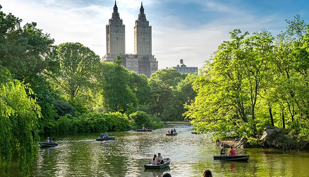 People are leisurely rowing boats on a tranquil pond surrounded by lush greenery with a pair of towering residential buildings in the background giving a serene juxtaposition of nature and urban architecture