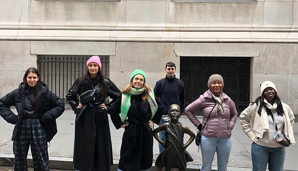 A group of people is standing around the Fearless Girl statue each adopting a similar pose with their hands on their hips emulating the statues confident stance