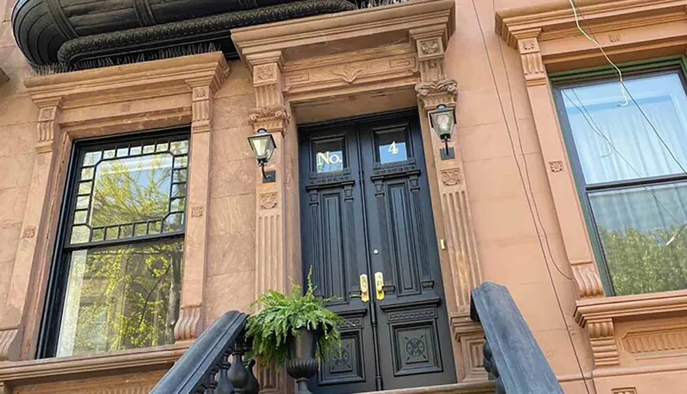 The image shows an elegant black double-door entrance to a building marked with the number 4 flanked by classical brownstone architecture and ornate details including a hanging fern under bright daylight