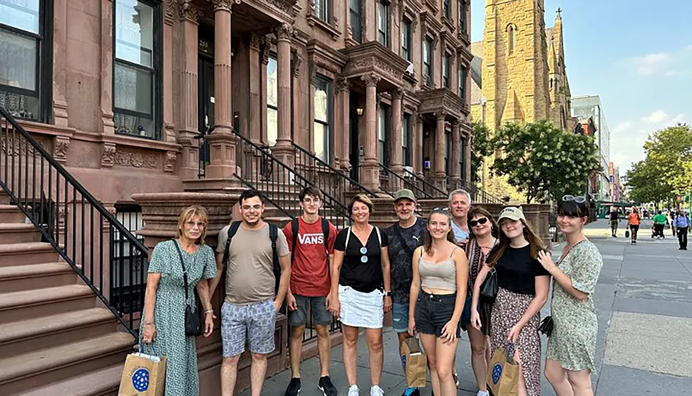 A group of smiling people poses for a photo on the sidewalk in front of a row of brownstone buildings indicative of an urban residential neighborhood