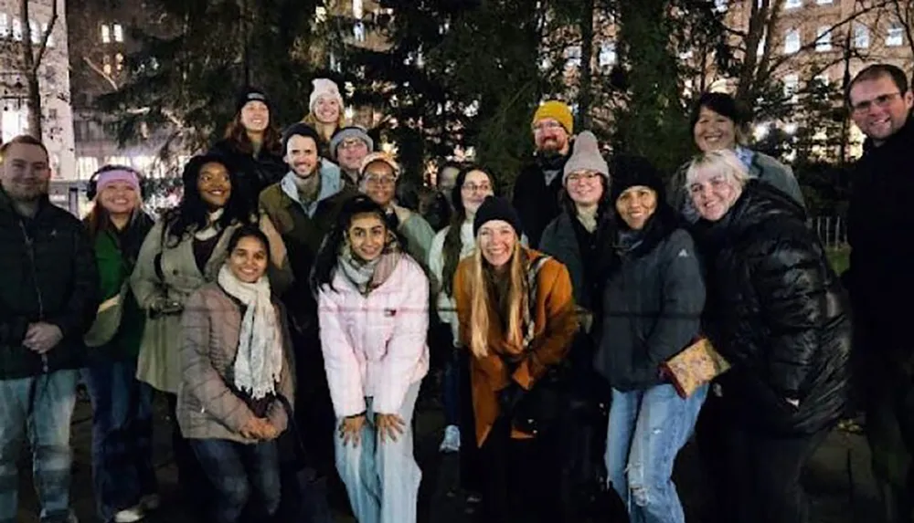 A group of smiling people pose for a photo at an outdoor evening gathering wearing winter clothing