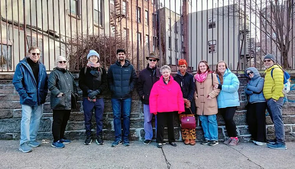 A diverse group of people are posing for a photo outdoors on steps with a backdrop of a metal fence and brick buildings