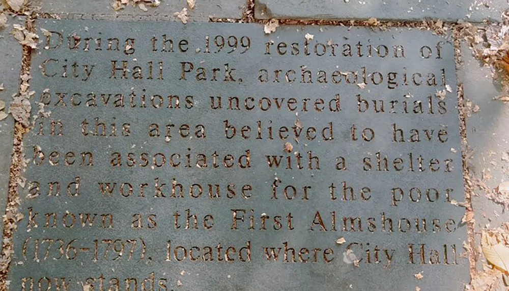 The image shows a weathered informational plaque detailing that archaeological excavations during the 1999 restoration of City Hall Park uncovered burials in an area once occupied by the First Almshouse a shelter and workhouse for the poor