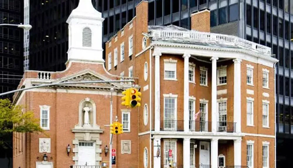 The image shows a historic brick building with white trim and a classical design standing in stark contrast to the modern glass and steel skyscraper that looms behind it