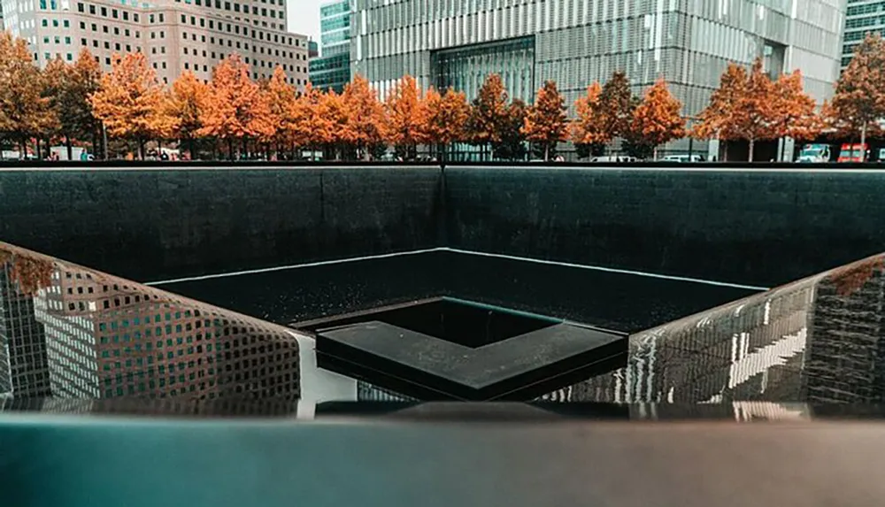 The image shows one of the 911 Memorial pools surrounded by trees with autumn foliage against a backdrop of modern buildings