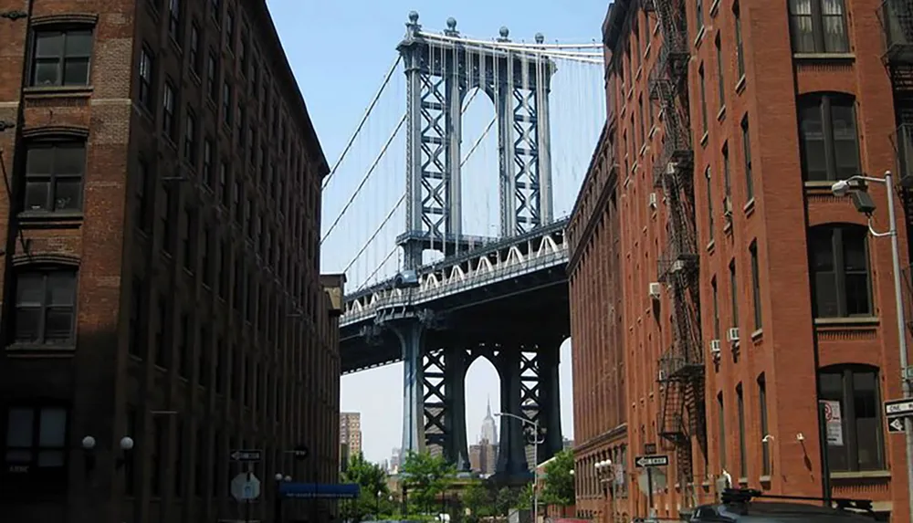 A view from a street framed by old brick buildings leading the eye to the towering structure of a suspension bridge under a clear sky