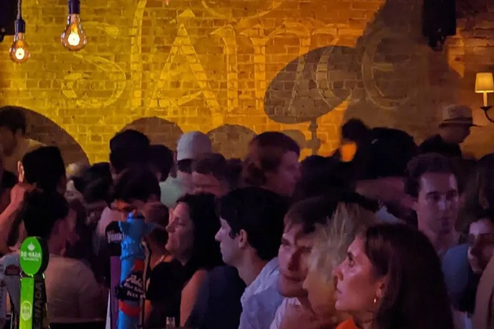 The image shows a busy and dimly lit bar scene with several patrons gathered some looking towards the camera under warm ambient lighting