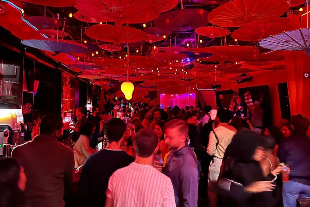 The image depicts a bustling bar or nightclub scene with a vibrant atmosphere characterized by red lighting and numerous paper umbrellas suspended from the ceiling