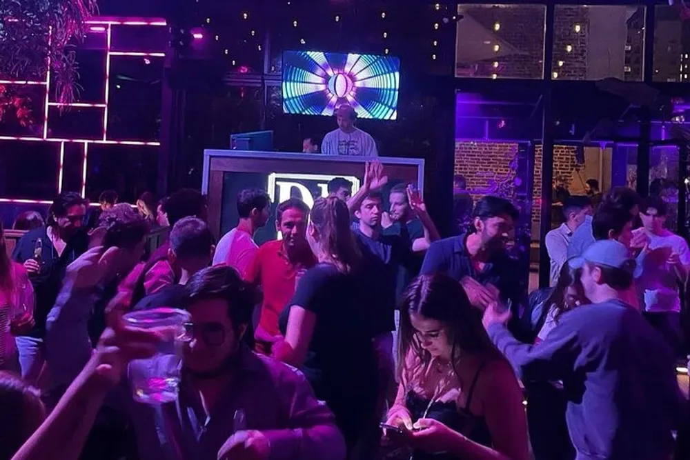 The image shows a lively night scene at a club with a DJ performing in the background and a crowd of people dancing and socializing