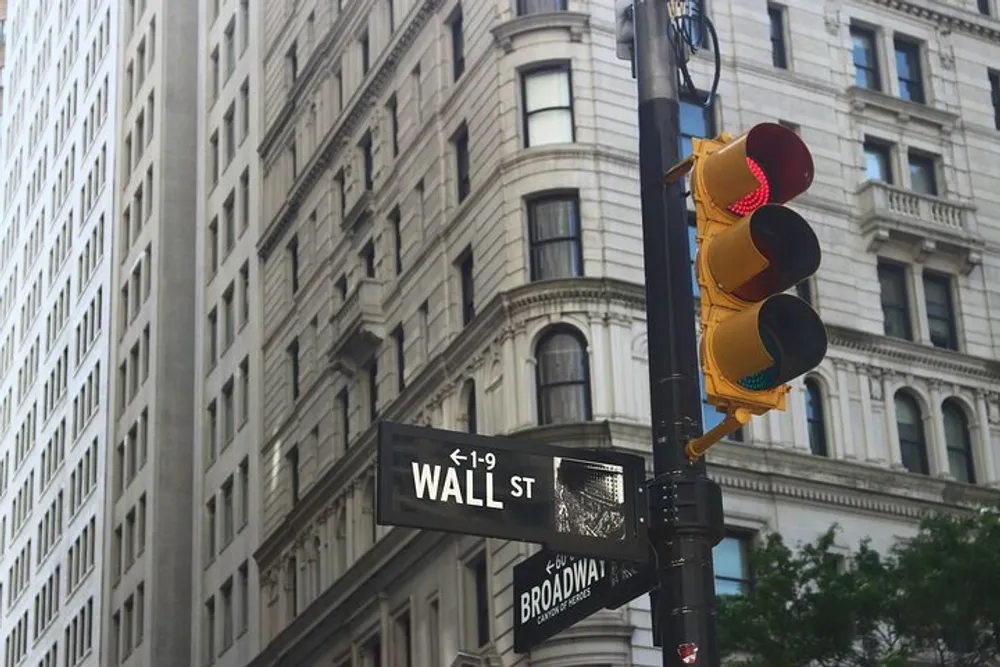 A red traffic light hangs above the street signs for Wall Street and Broadway at an intersection with classic architecture in the background