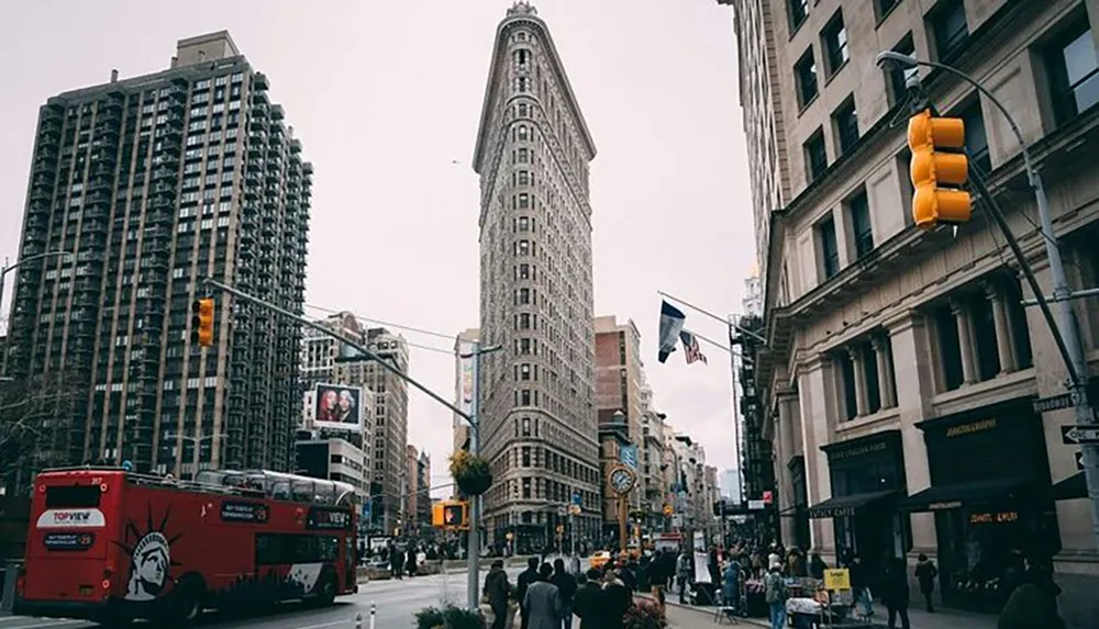 The image shows a bustling city street with pedestrians and a red double-decker tour bus in front of a distinctive triangular flatiron building