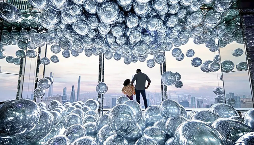 Two individuals stand amidst a cluster of reflective spheres gazing out at an urban skyline from a high vantage point