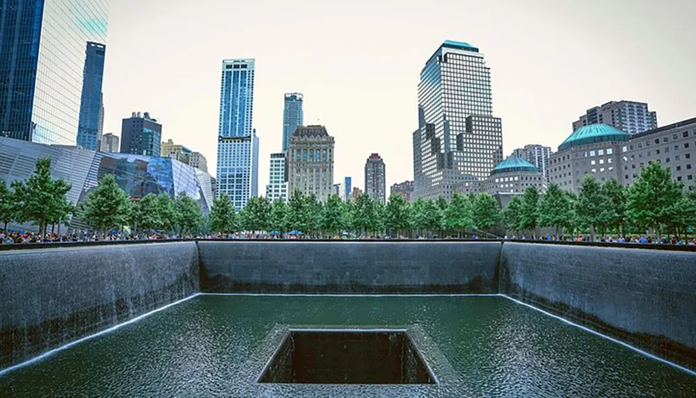 This image shows one of the 911 Memorial pools in New York City with cascading waterfalls leading into a square void surrounded by trees and skyscrapers in the background