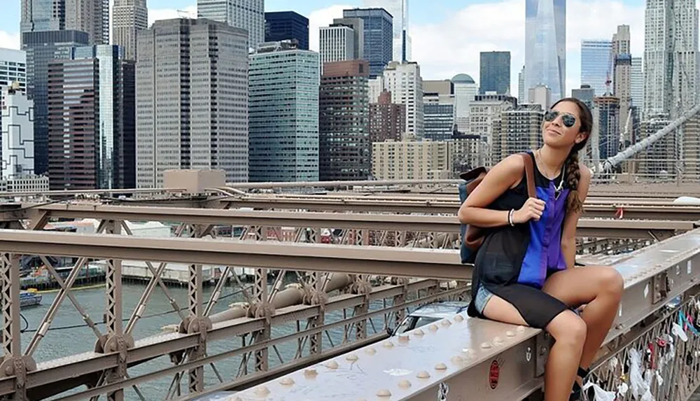 A person is smiling while sitting on the Brooklyn Bridge with the New York City skyline in the background