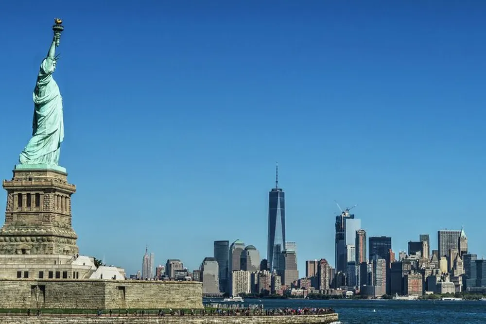 The Statue of Liberty stands prominently in the foreground with the Manhattan skyline and One World Trade Center in the background under a clear blue sky