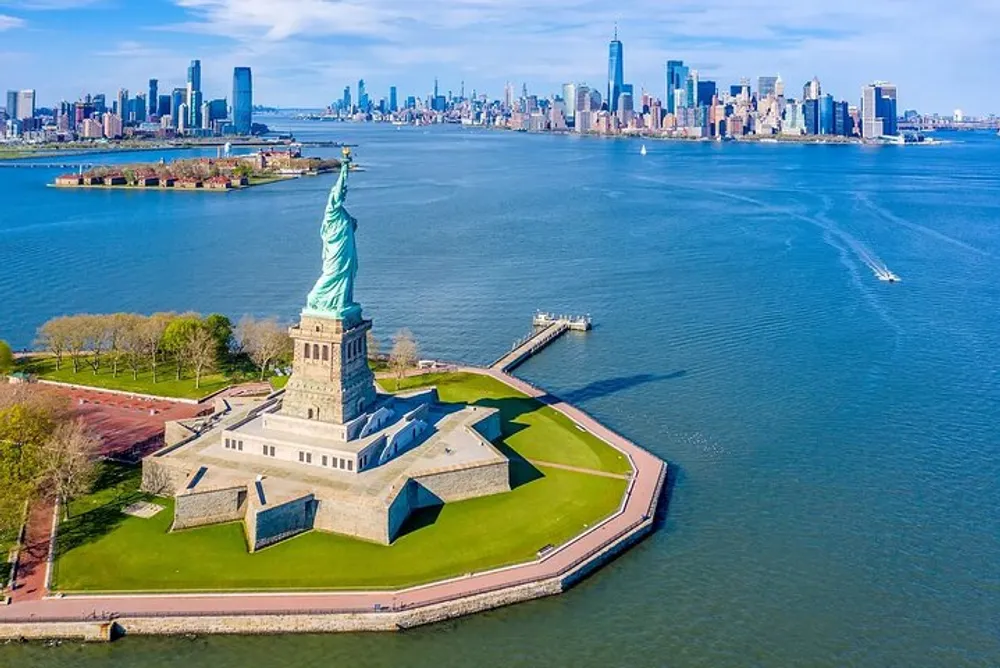 The image shows a sunny aerial view of the Statue of Liberty with a backdrop of New York Citys skyline and a clear blue sky