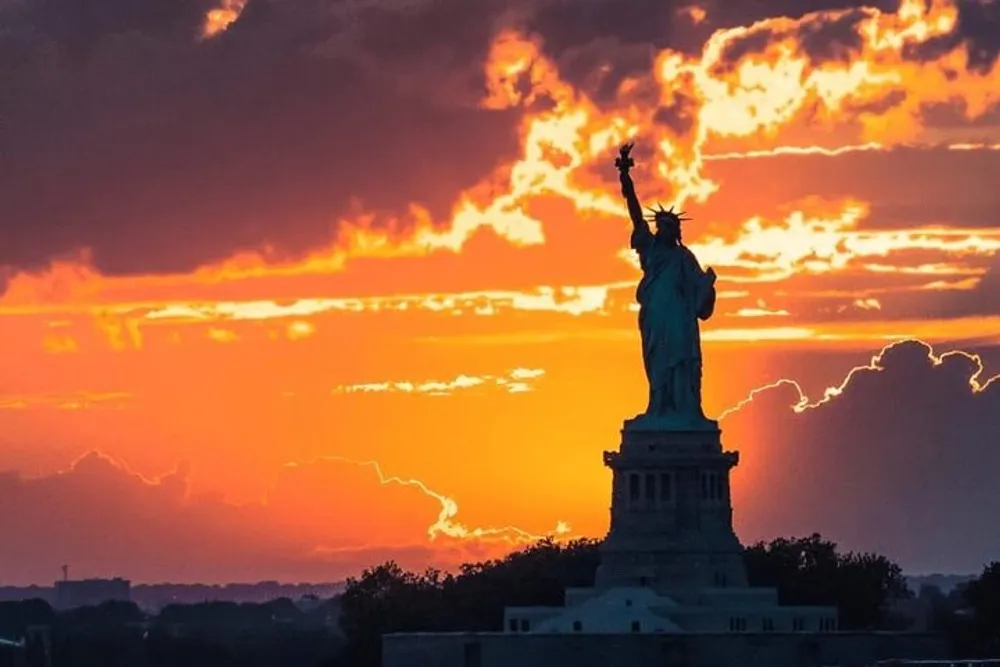 The Statue of Liberty is silhouetted against a dramatic orange and yellow sunset sky with visible clouds and light rays