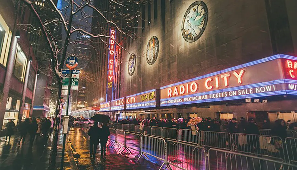 Pedestrians with umbrellas walk past the neon-lit faade of Radio City Music Hall on a rainy evening