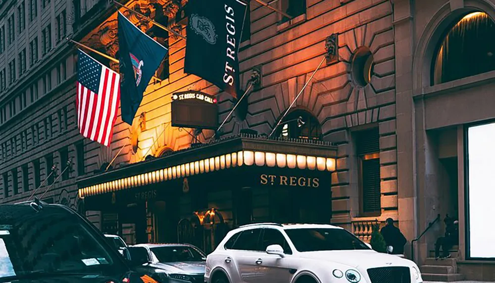 The image shows the lit entrance of the St Regis hotel with multiple flags flying above including the American flag amidst parked cars and a person walking by the building